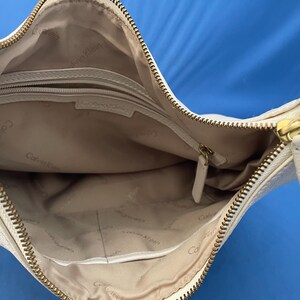 May include: Cream-colored Calvin Klein crossbody bag with a gold zipper and hardware. The interior is beige with a zippered pocket and the Calvin Klein logo. The bag is open, showing the inside.