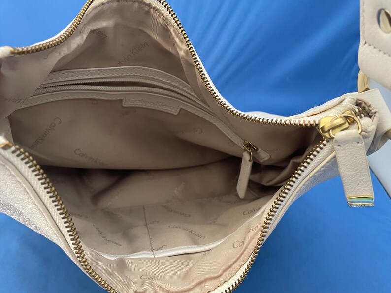 May include: Cream-colored Calvin Klein crossbody bag with a gold zipper and hardware. The interior is beige with a zippered pocket and the Calvin Klein logo. The bag is open, showing the inside.
