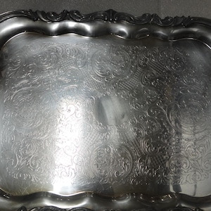 Op de afbeelding: Een zilveren dienblad met sierlijke handvatten en een decoratief reliëfpatroon. Het dienblad heeft een licht gegolfde rand.