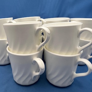 Può includere: Una collezione di tazze in ceramica bianca disposte in gruppo. Ogni tazza presenta una finitura liscia e lucida con un sottile motivo a spirale diagonale impresso sulla superficie esterna. Le tazze hanno un manico curvo e un bordo circolare.