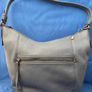 May include: A light blue leather shoulder bag with a curved shape. The bag has a zipper closure and a long, rounded strap. Additional features include a front zippered pocket and a small metal charm.