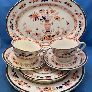 May include: A ceramic dinnerware set featuring an oval platter, two teacups with matching saucers, and two dinner plates. Each piece is white with a decorative pattern of orange and dark blue flowers, accented by thin gold trim along the edges.