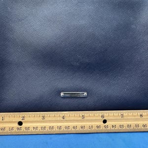 Puede incluir: Un bolso bandolera azul marino con una correa de cadena plateada. El bolso tiene una superficie texturizada y un peque&ntilde;o logotipo rectangular plateado. Una regla de madera se coloca debajo, midiendo hasta 23 cm.