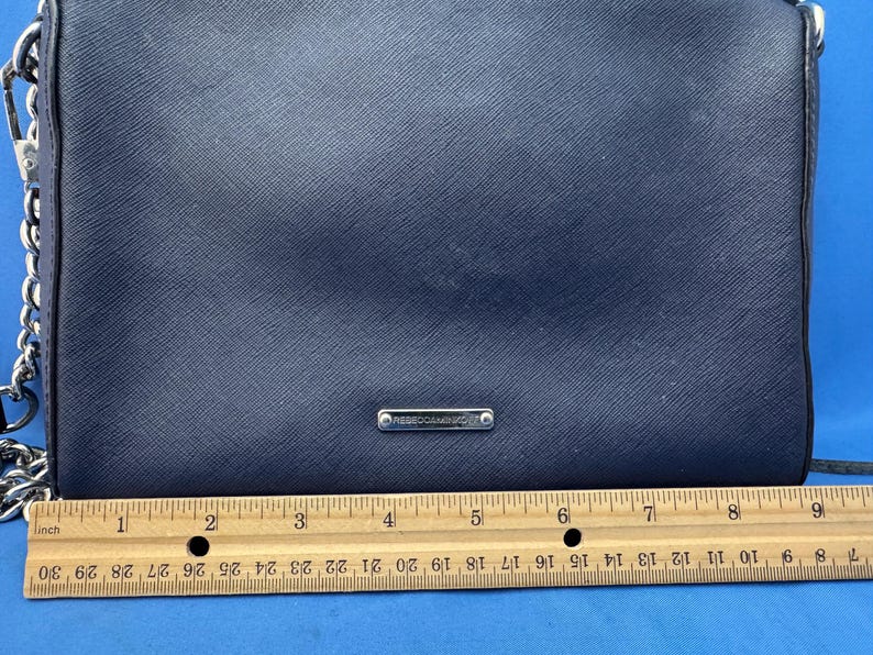 Puede incluir: Un bolso bandolera azul marino con una correa de cadena plateada. El bolso tiene una superficie texturizada y un peque&ntilde;o logotipo rectangular plateado. Una regla de madera se coloca debajo, midiendo hasta 23 cm.