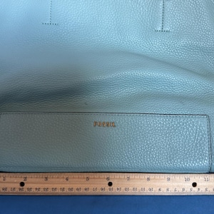 May include: A light blue Fossil tote bag with a textured leather appearance. The bag has a rectangular front pocket with the brand name "FOSSIL" in gold lettering. A ruler is placed below the bag, measuring approximately 12 inches.