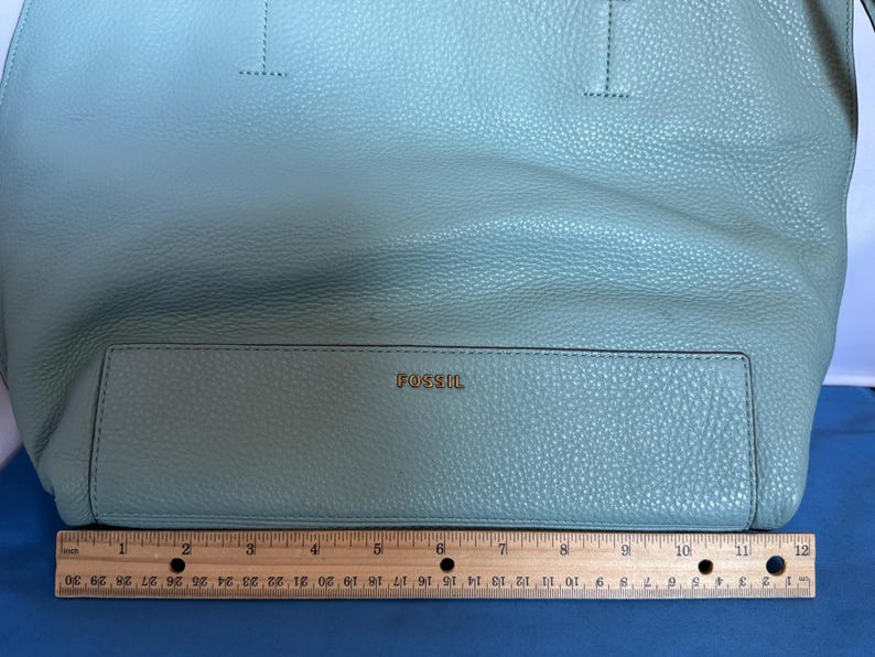 May include: A light blue Fossil tote bag with a textured leather appearance. The bag has a rectangular front pocket with the brand name "FOSSIL" in gold lettering. A ruler is placed below the bag, measuring approximately 12 inches.