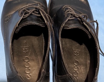 May include: A pair of black leather dress shoes with laces. The shoes have a rounded toe and a low heel. The brand name "ECCO" is visible inside the shoe. The shoes are on a blue surface.