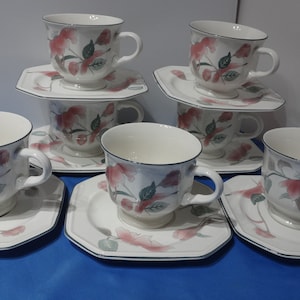May include: Set of six white teacups and saucers with a floral design featuring pink flowers and green leaves. The cups and saucers have a black trim.