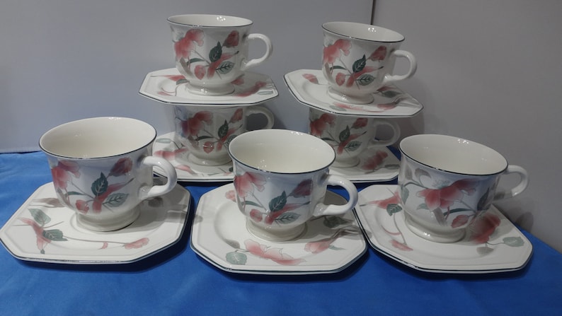 May include: Set of six white teacups and saucers with a floral design featuring pink flowers and green leaves. The cups and saucers have a black trim.
