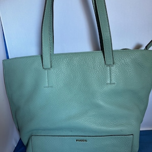 May include: A light teal leather tote bag with long handles. The bag has a front pocket and the brand name "FOSSIL" is embossed on the front. The bag is made of textured leather.