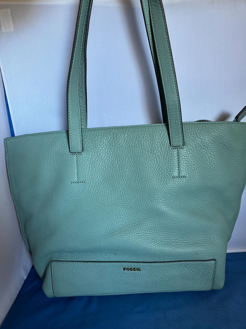 May include: A light teal leather tote bag with long handles. The bag has a front pocket and the brand name "FOSSIL" is embossed on the front. The bag is made of textured leather.