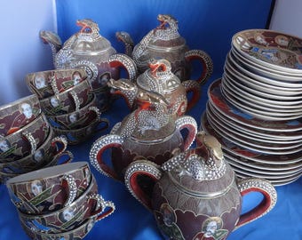 May include: A vintage Japanese tea set with a red and gold floral design. The set includes a teapot, sugar bowl, creamer, and six cups and saucers. The set is decorated with intricate details and features a traditional Japanese design.