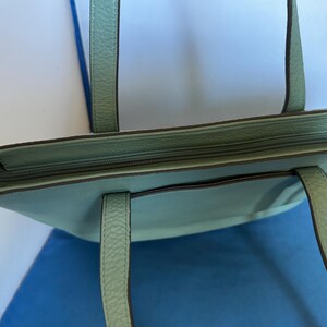May include: A light teal tote bag with a zipper closure and two handles. The bag has a textured surface and a small pocket on the front. The handles are a matching teal color. The bag is on a blue surface.