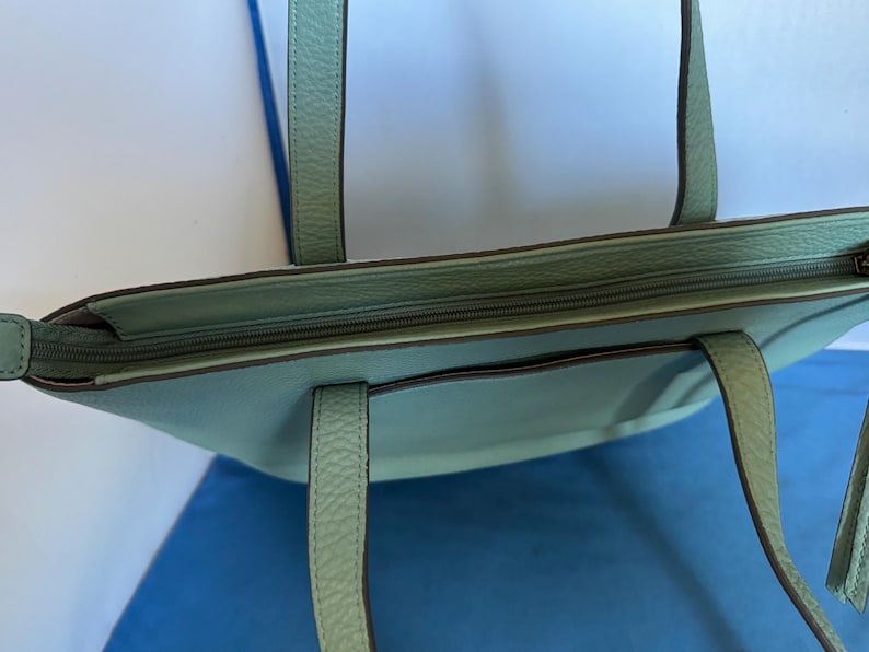 May include: A light teal tote bag with a zipper closure and two handles. The bag has a textured surface and a small pocket on the front. The handles are a matching teal color. The bag is on a blue surface.
