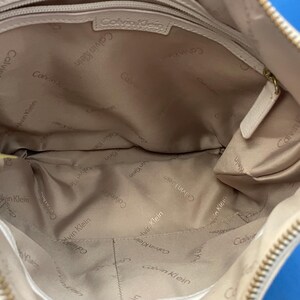 May include: Beige Calvin Klein handbag. The interior is lined with a light tan fabric, with the Calvin Klein logo printed repeatedly. The bag has a zipper closure and a small pocket inside. The bag is made of a soft, smooth material.