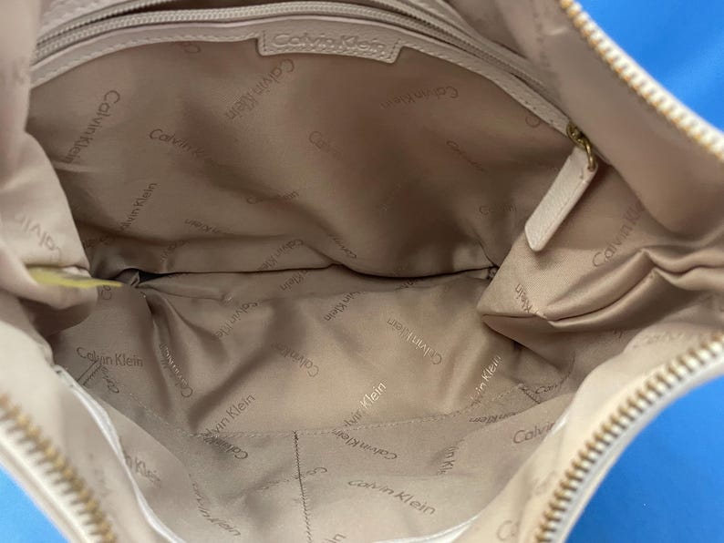 May include: Beige Calvin Klein handbag. The interior is lined with a light tan fabric, with the Calvin Klein logo printed repeatedly. The bag has a zipper closure and a small pocket inside. The bag is made of a soft, smooth material.