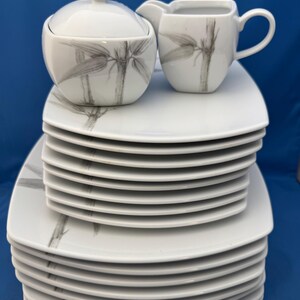 Może przedstawiać: Biały zestaw porcelanowej zastawy stołowej z motywem bambusa. Zestaw zawiera cukiernicę z pokrywką, dzbanuszek do śmietanki i stos kwadratowych talerzy. Wzór bambusa jest w odcieniach szarości. Na niebieskim tle.