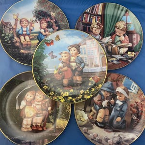 Op de afbeelding: Set van vijf decoratieve borden met illustraties van kinderen in traditionele Duitse kleding. Elk bord toont een andere scène in een charmante, grillige stijl. De borden zijn wit met gouden rand en dragen de handtekening van de kunstenaar.