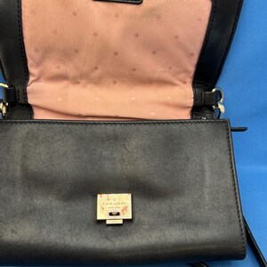 Może przedstawiać: Czarna sk&oacute;rzana torebka crossbody o prostokątnym kształcie. Torebka posiada złoty zatrzask i okucia. Wnętrze jest w kolorze jasnor&oacute;żowym z subtelnym wzorem w kropki. Nazwa marki "Kate Spade New York" jest widoczna na zatrzasku.