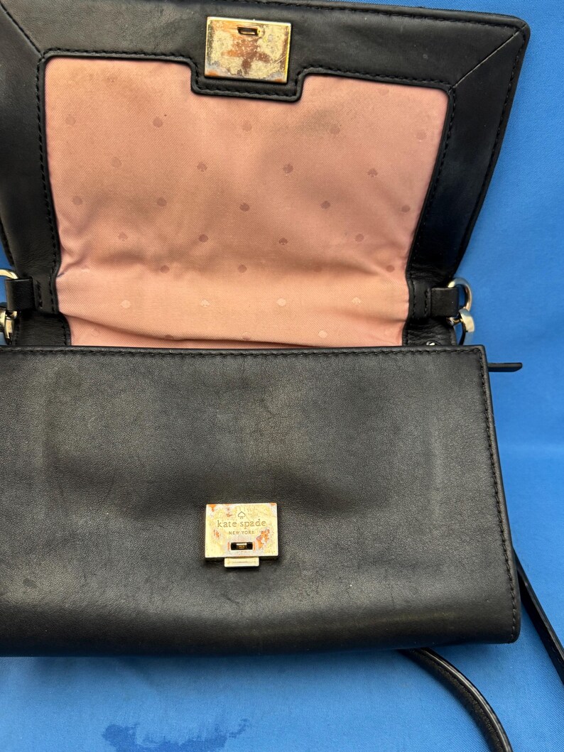 Może przedstawiać: Czarna sk&oacute;rzana torebka crossbody o prostokątnym kształcie. Torebka posiada złoty zatrzask i okucia. Wnętrze jest w kolorze jasnor&oacute;żowym z subtelnym wzorem w kropki. Nazwa marki "Kate Spade New York" jest widoczna na zatrzasku.