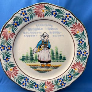Könnte beinhalten: Dekorative Keramikplatte mit gewelltem Rand, mit handgemaltem Design. Das zentrale Bild zeigt eine Frau in traditioneller Kleidung, umgeben von einem floralen Rand mit roten, blauen und grünen Akzenten. Die Platte hat einen braunen Rand und einen goldenen Rand.