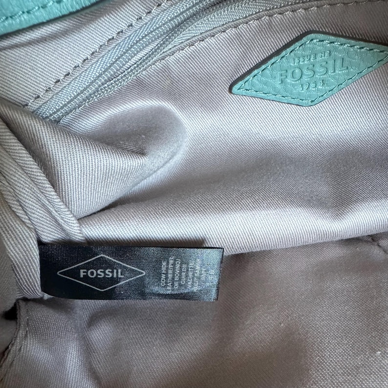 May include: Interior view of a Fossil handbag, showing the lining and branding. The bag's interior is a light gray fabric. A teal leather diamond-shaped tag with the Fossil logo is visible. A black tag with the Fossil logo and text is also present.