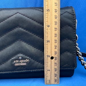 Może przedstawiać: Czarny pikowany sk&oacute;rzany portfel ze srebrnym łańcuszkiem. Portfel ma wz&oacute;r w szewron i srebrne logo "kate spade NEW YORK". Portfel ma około 25 cm długości.