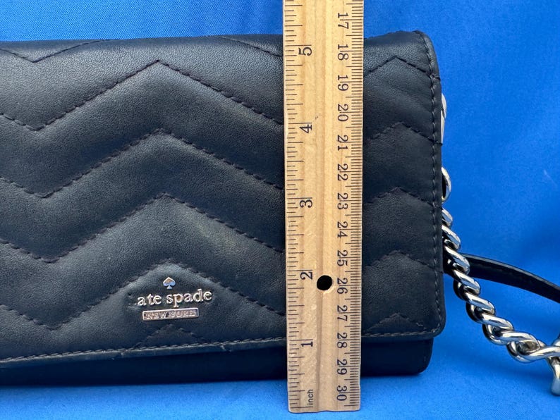 Może przedstawiać: Czarny pikowany sk&oacute;rzany portfel ze srebrnym łańcuszkiem. Portfel ma wz&oacute;r w szewron i srebrne logo "kate spade NEW YORK". Portfel ma około 25 cm długości.