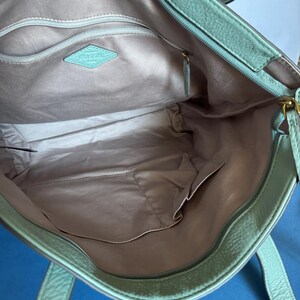 May include: Open view of a mint green leather tote bag. The interior features a light gray lining with multiple pockets and a zippered compartment. A diamond-shaped tag with the brand name is visible inside the bag.