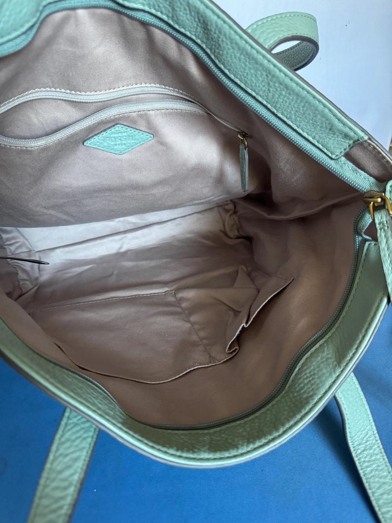 May include: Open view of a mint green leather tote bag. The interior features a light gray lining with multiple pockets and a zippered compartment. A diamond-shaped tag with the brand name is visible inside the bag.