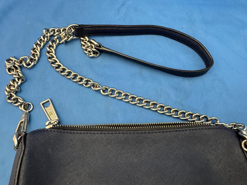 Puede incluir: Un bolso cruzado azul marino con una correa de cadena plateada y un asa negra. El bolso tiene un cierre de cremallera y una superficie texturizada. La cadena est&aacute; hecha de eslabones grandes e interconectados.