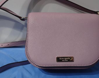 Może przedstawiać: Jasno-liliowa skórzana torebka na ramię z złotą klamrą i napisem "kate spade new york" z przodu.
