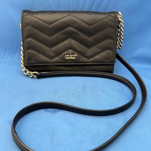 Może przedstawiać: Czarna pikowana torebka crossbody z wzorem w jodełkę. Torba posiada złoty łańcuszek i długi, cienki czarny pasek. Logo Kate Spade jest widoczne z przodu torby.