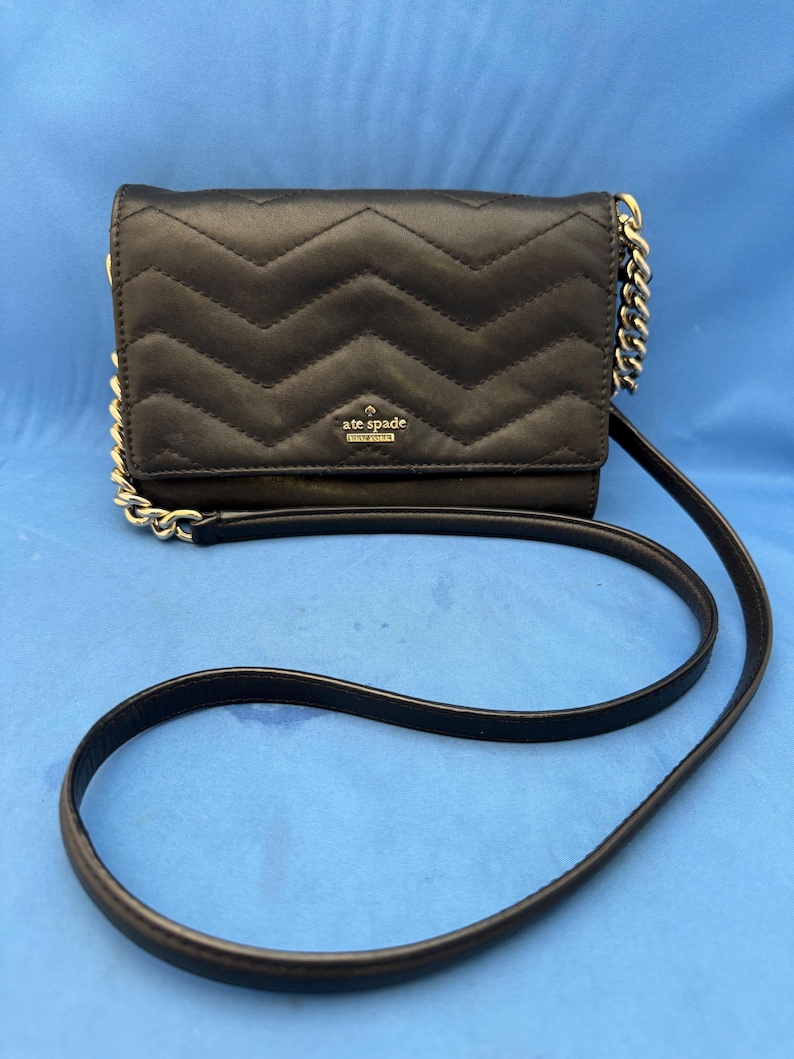Może przedstawiać: Czarna pikowana torebka crossbody z wzorem w jodełkę. Torba posiada złoty łańcuszek i długi, cienki czarny pasek. Logo Kate Spade jest widoczne z przodu torby.