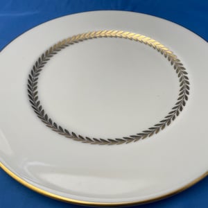 May include: A white ceramic dinner plate with a gold rim and a black and gold leaf design around the edge.