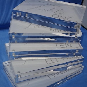 May include: A stack of five clear acrylic table number holders with the numbers one, nine, ten, eleven, and twelve printed on them in a gray font.
