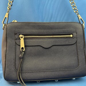 Puede incluir: Un bolso de mano azul marino con una cadena dorada y cremallera dorada. El bolso tiene forma rectangular con un bolsillo frontal y un tirador dorado. El bolso est&aacute; hecho de un material texturizado y tiene un dise&ntilde;o minimalista.