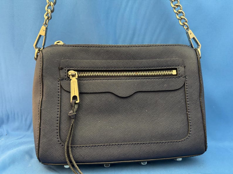 Puede incluir: Un bolso de mano azul marino con una cadena dorada y cremallera dorada. El bolso tiene forma rectangular con un bolsillo frontal y un tirador dorado. El bolso est&aacute; hecho de un material texturizado y tiene un dise&ntilde;o minimalista.