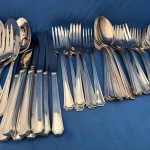 May include: An assortment of silver-coloured cutlery, including spoons, forks, and knives, arranged on a blue surface. The silverware features a classic design with decorative handles. The image showcases a variety of dining utensils.