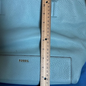 May include: A light blue Fossil tote bag. The bag is made of textured leather and has two handles. The bag is approximately 12 inches tall. The brand name "FOSSIL" is visible on the front of the bag.