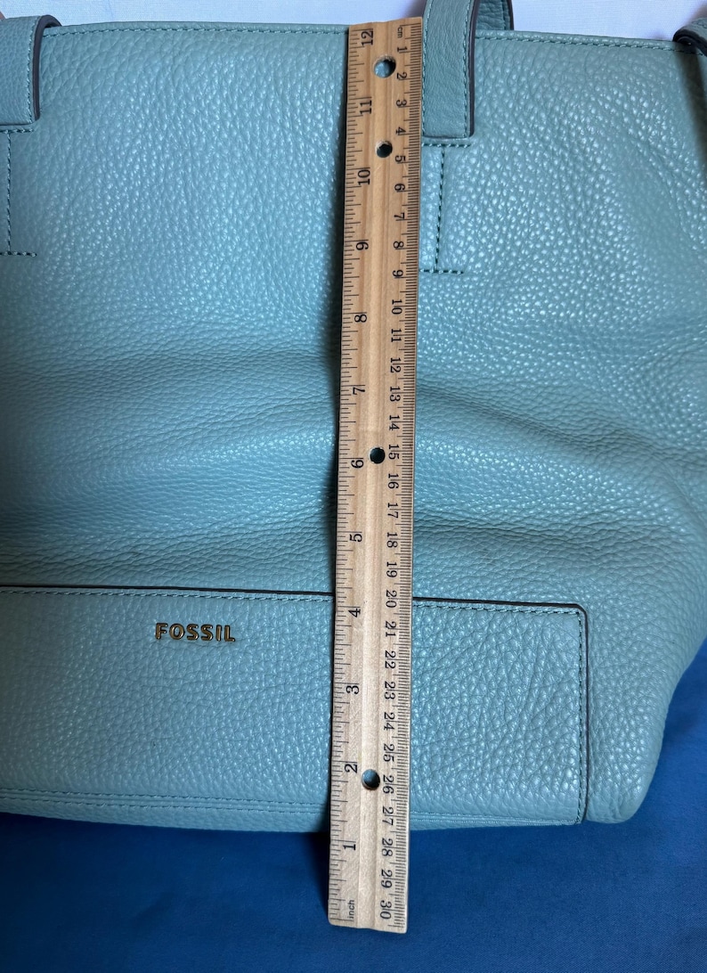 May include: A light blue Fossil tote bag. The bag is made of textured leather and has two handles. The bag is approximately 12 inches tall. The brand name "FOSSIL" is visible on the front of the bag.