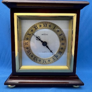 May include: A rectangular, dark brown wooden clock with a gold-colored square frame around the clock face. The clock face is cream-colored with black Roman numerals and the word "ANGELUS" above the hands. The clock is a quartz movement.