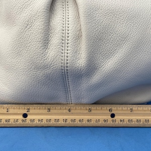May include: Off-white leather handbag with a textured surface. The bag is partially visible, with a stitched seam detail. A wooden ruler is placed below the bag, showing measurements in inches.