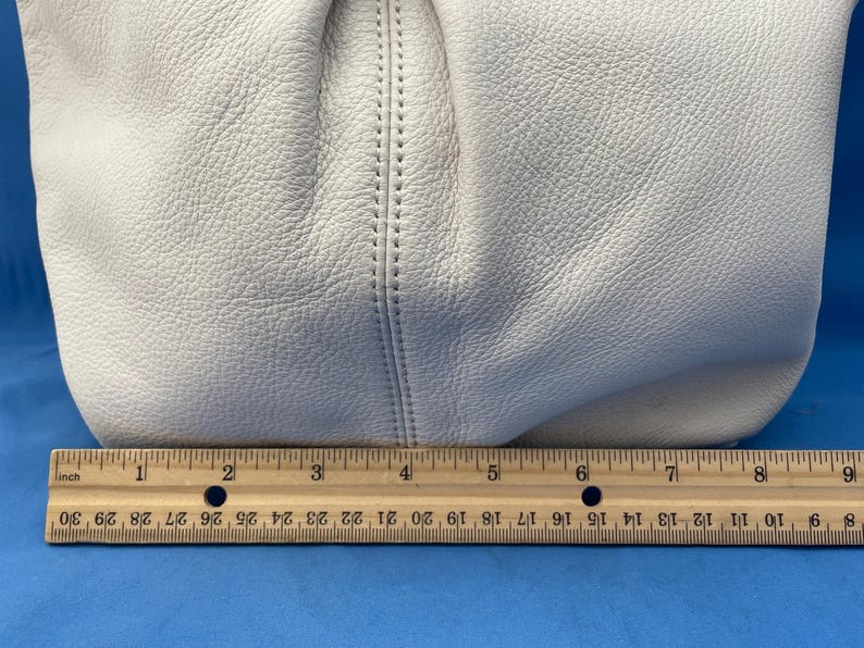 May include: Off-white leather handbag with a textured surface. The bag is partially visible, with a stitched seam detail. A wooden ruler is placed below the bag, showing measurements in inches.
