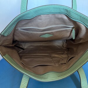 May include: Open view of a mint green leather tote bag. The interior features a light brown lining with multiple pockets, including a zippered compartment. The bag has two handles and a gold-colored zipper pull.