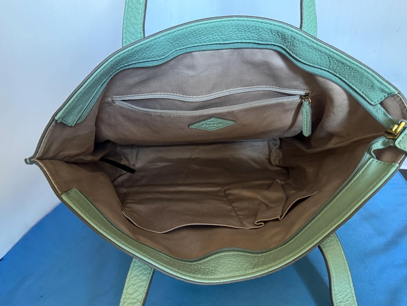 May include: Open view of a mint green leather tote bag. The interior features a light brown lining with multiple pockets, including a zippered compartment. The bag has two handles and a gold-colored zipper pull.
