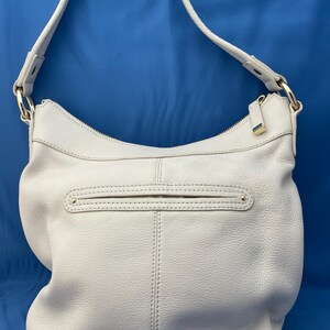 May include: A light gray leather shoulder bag with a long strap. The bag has a curved shape with a zippered top and a front pocket. Gold-colored hardware accents the bag, including the zipper pulls and strap attachments.