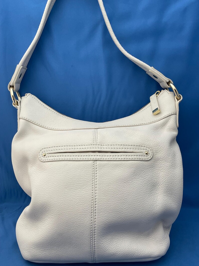 May include: A light gray leather shoulder bag with a long strap. The bag has a curved shape with a zippered top and a front pocket. Gold-colored hardware accents the bag, including the zipper pulls and strap attachments.