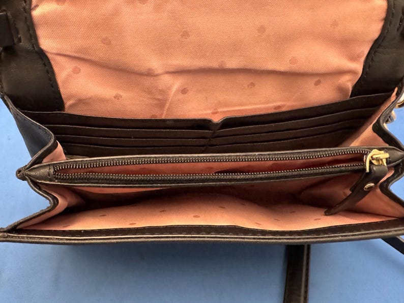 Może przedstawiać: Czarna sk&oacute;rzana torba crossbody, otwarta, aby odsłonić wiele przegr&oacute;dek na karty i przegrodę zapinaną na zamek. Wnętrze jest w kolorze jasnor&oacute;żowym z wzorem w kropki. Torba ma złoty zamek błyskawiczny i okucia.