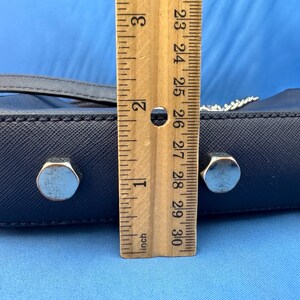 Puede incluir: Un bolso de mano azul marino con herrajes plateados. El bolso mide aproximadamente 5 cm de altura. Se coloca una regla de madera junto al bolso para medir.
